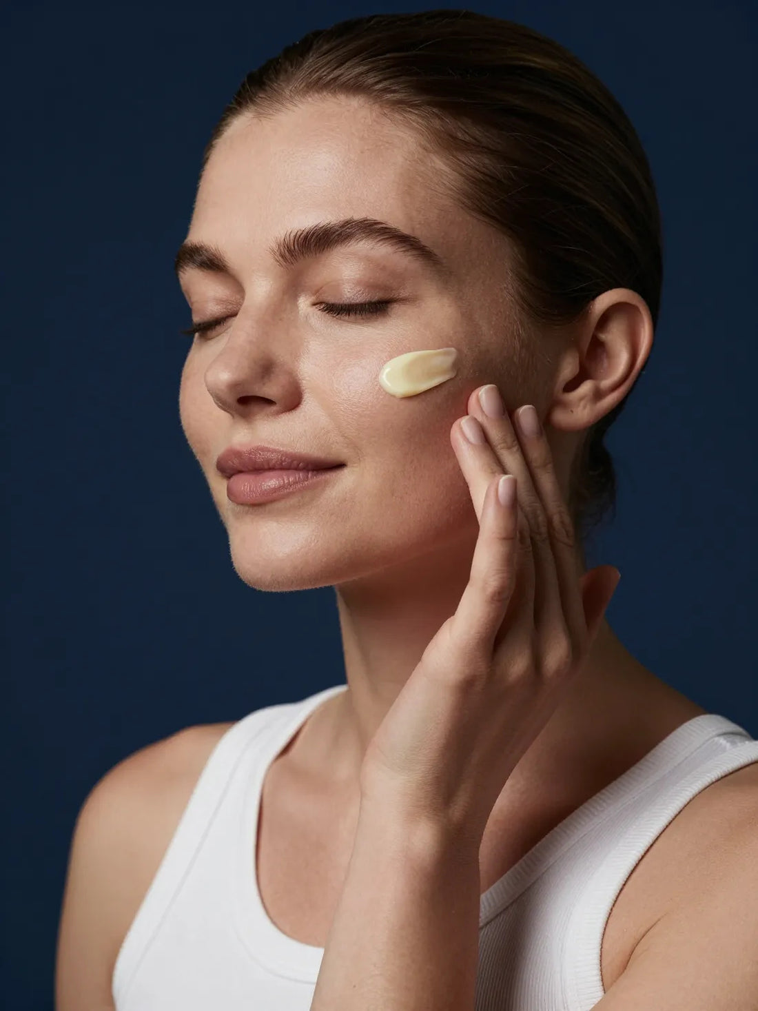 A pale yellow serum pressed into the cheek with the fingertips, eyes closed, CELLULAR ANTI-AGING ASCORBYL GLUCOSIDE SERUM