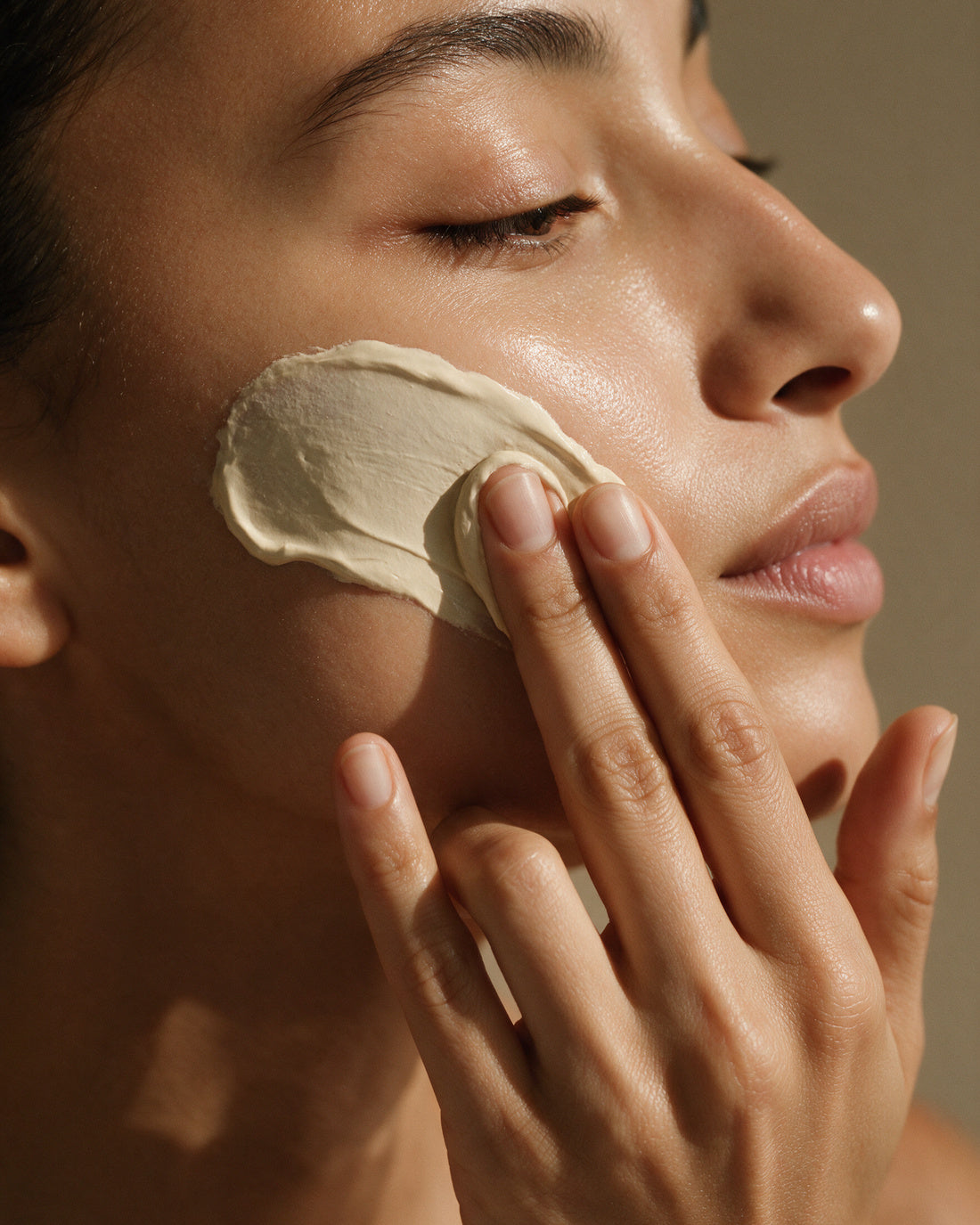 A smooth pale clay mask being smoothed across the cheekbone