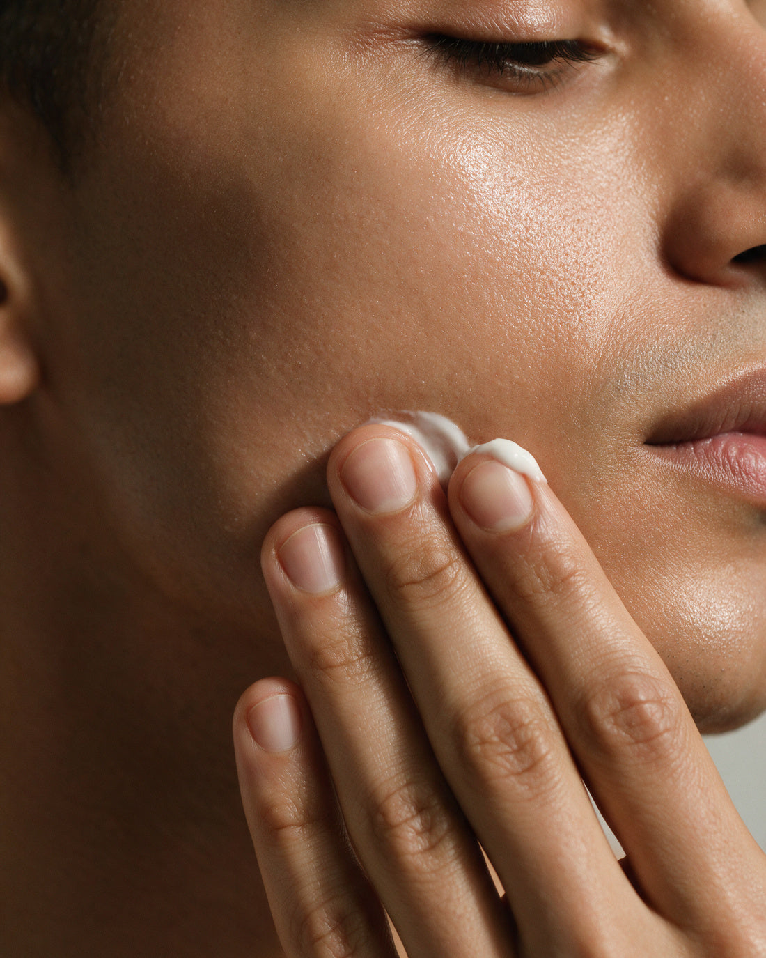 A man's jaw and cheek, fingertips pressing a small dab of white cream into the skin